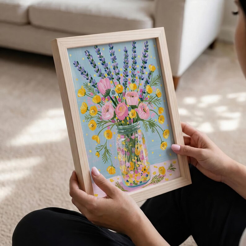 May include: A framed floral print featuring a bouquet of pink and yellow flowers, lavender, and greenery in a mason jar. The artwork has a light blue background and is set in a natural wood frame. The painting is held by a person.