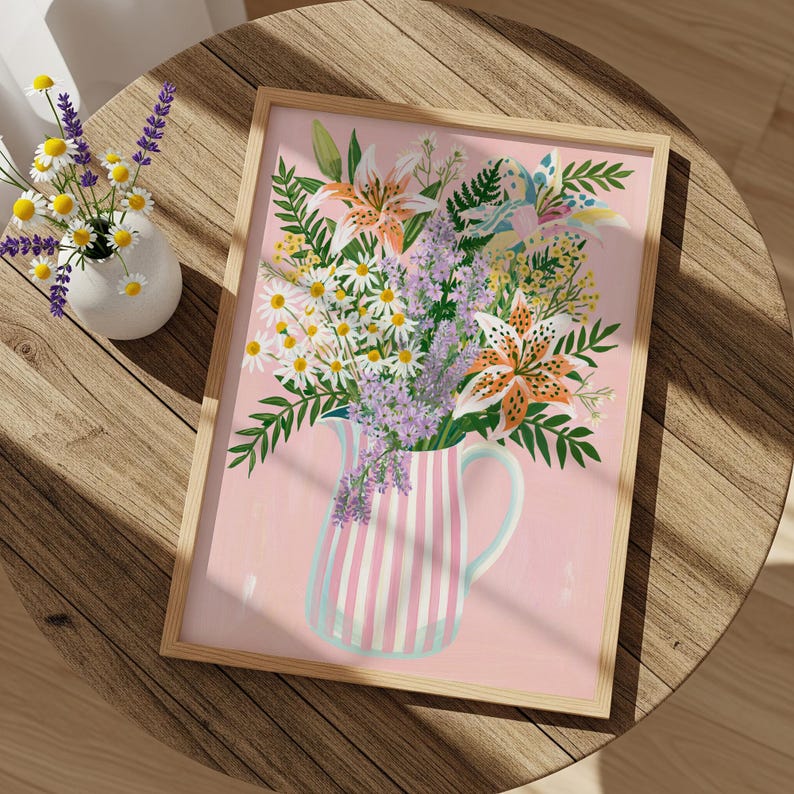 Pu&ograve; includere: Una stampa floreale incorniciata con un bouquet di gigli, margherite e lavanda in una brocca a righe rosa e bianche. L'opera d'arte &egrave; ambientata su uno sfondo rosa tenue e incorniciata in legno naturale. Un piccolo vaso di fiori &egrave; vicino.