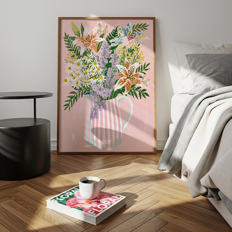 Pu&ograve; includere: Una stampa artistica incorniciata raffigurante un bouquet di fiori, tra cui gigli e margherite, in una brocca a righe rosa e bianche. L'opera d'arte &egrave; ambientata su uno sfondo rosa. Una tazza di caff&egrave; &egrave; appoggiata su una rivista con la parola "FLORAL".