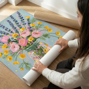 May include: A floral art print featuring a bouquet of pink roses, yellow flowers, and purple lavender in a glass jar against a light blue background. A rolled-up white canvas is held by a person.