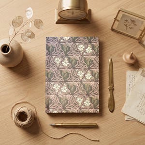 May include: A floral patterned journal with a gold pen, letter opener, and clock on a wooden surface. The journal features a repeating pattern of white flowers and green leaves on a mauve background. A small vase with a plant and a spool of twine are also present.