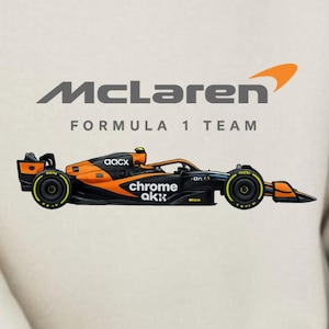 Può includere: Felpa color crema con il logo McLaren Formula 1 Team e una grafica di un'auto da corsa nera, arancione e gialla. Il testo "McLaren Formula 1 Team" è stampato sopra l'auto.