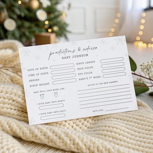 May include: A white baby shower advice card with the text "predictions & advice" and "Baby Johnson". The card has spaces for birth details and advice, with snowflake accents. It rests on a cream-coloured knitted blanket.