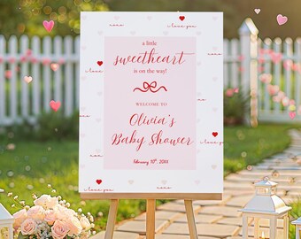 May include: A baby shower sign on a wooden easel, with the text "a little sweetheart is on the way!" in red script. The sign is surrounded by white lanterns, flowers, and a picket fence.