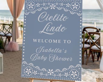 May include: A blue sign on a wooden easel with the text "Cielito Lindo Welcome to Isabella's Baby Shower." The sign has a white floral border and is set up at an outdoor event with chairs and decorations.