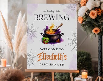 May include: A sign on an easel for a baby shower, with the text "a baby is BREWING" and "Welcome to Elizabeth's Baby Shower." The sign features a cauldron graphic with a skull and spiderwebs. Pumpkins and flowers decorate the scene.