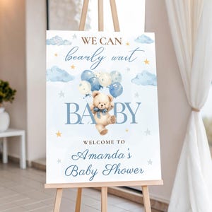 May include: A baby shower welcome sign on an easel. The sign features a teddy bear holding balloons, with the words "We can bearly wait BABY" and "Welcome to Amanda's Baby Shower" in blue script.