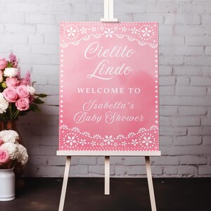May include: A pink baby shower welcome sign on a white easel. The sign reads "Cielito Lindo, Welcome to Isabella's Baby Shower" in white script. Floral decorations frame the text. A bouquet of pink roses sits nearby.