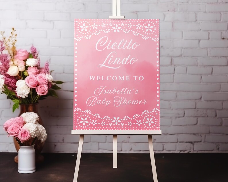 May include: A pink baby shower welcome sign on a white easel. The sign reads "Cielito Lindo, Welcome to Isabella's Baby Shower" in white script. Floral decorations frame the text. A bouquet of pink roses sits nearby.