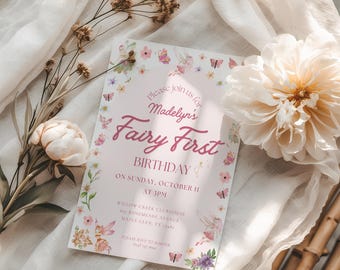 May include: A pink invitation card with the text "Madelyn's Fairy First Birthday" surrounded by floral and butterfly illustrations. The card is on a white fabric with a flower and dried floral arrangement.