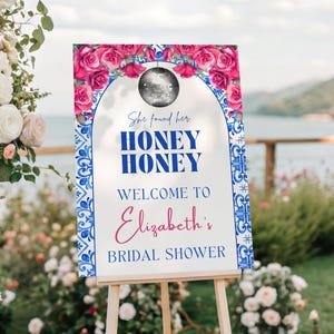May include: A welcome sign for a bridal shower, displayed on an easel. The sign features a floral design with pink roses and blue accents. Text reads "She found her HONEY HONEY Welcome to Elizabeth's Bridal Shower."