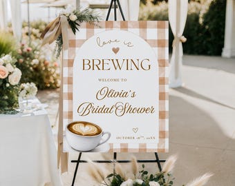 May include: A sign on an easel with a brown and white checkered background. The sign reads "Love is Brewing" and welcomes guests to Olivia's Bridal Shower. A coffee cup with latte art is featured.