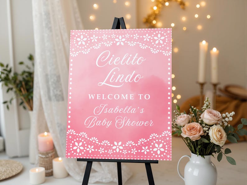 May include: A pink baby shower welcome sign on a black easel. The sign has white floral lace borders and text that reads "Cielito Lindo Welcome to Isabella's Baby Shower."