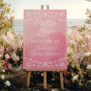 May include: A pink sign on an easel with the words "Cielito Lindo" and "Welcome to Isabella's Baby Shower" in white script. The sign is decorated with a floral border and set against a backdrop of flowers and the ocean.