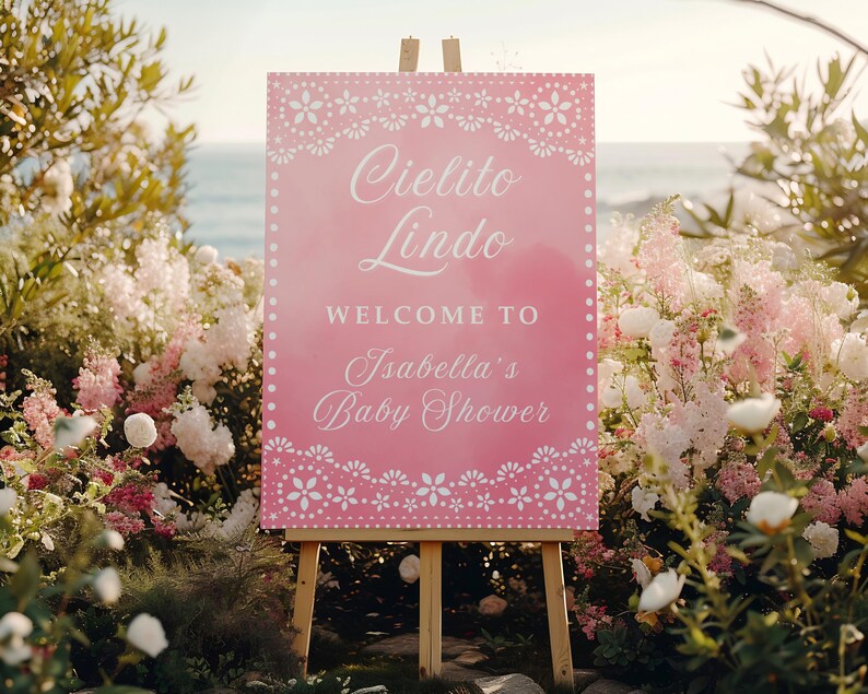 May include: A pink sign on an easel with the words "Cielito Lindo" and "Welcome to Isabella's Baby Shower" in white script. The sign is decorated with a floral border and set against a backdrop of flowers and the ocean.