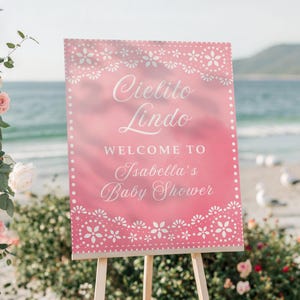 May include: A pink sign with white floral designs and the words "Cielito Lindo" and "Welcome to Isabella's Baby Shower." The sign is on a wooden easel, with flowers and the ocean in the background.