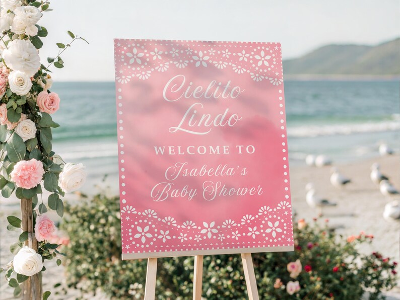 May include: A pink sign with white floral designs and the words "Cielito Lindo" and "Welcome to Isabella's Baby Shower." The sign is on a wooden easel, with flowers and the ocean in the background.