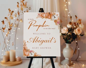 May include: A white sign on a black easel with the text "A little Pumpkin is on the way!" and "Welcome to Abigail's Baby Shower". The sign is decorated with pumpkins and floral arrangements in shades of orange, white, and pink.