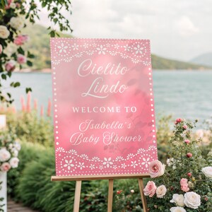 May include: A pink sign on a wooden easel with the words "Cielito Lindo" and "Welcome to Isabella's Baby Shower" in white script. The sign is decorated with a white floral border. The background includes flowers and a body of water.