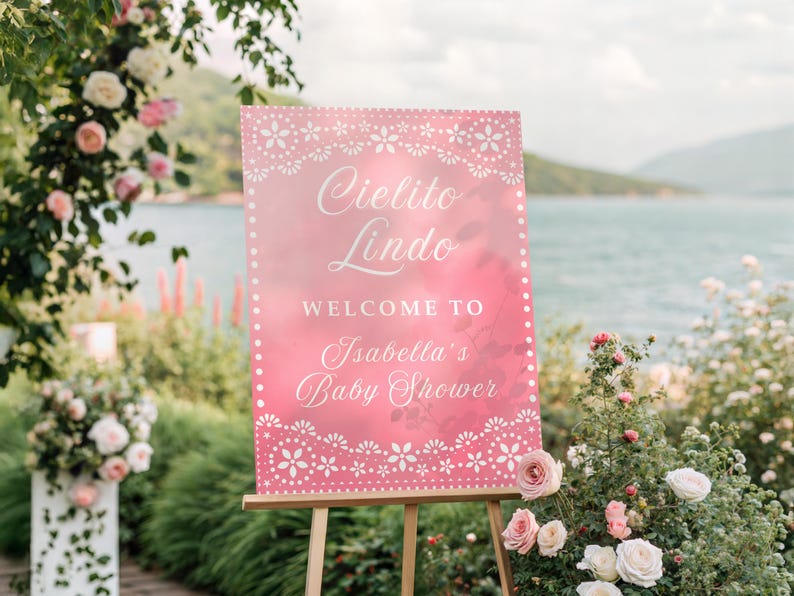 May include: A pink sign on a wooden easel with the words "Cielito Lindo" and "Welcome to Isabella's Baby Shower" in white script. The sign is decorated with a white floral border. The background includes flowers and a body of water.