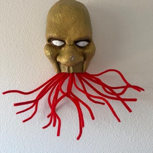 Puede incluir: Una m&aacute;scara dorada hecha a mano con ojos blancos y un dise&ntilde;o rojo en forma de tent&aacute;culos que se extiende desde la boca. La m&aacute;scara tiene una superficie texturizada y est&aacute; montada en una pared blanca, creando una exhibici&oacute;n art&iacute;stica &uacute;nica.