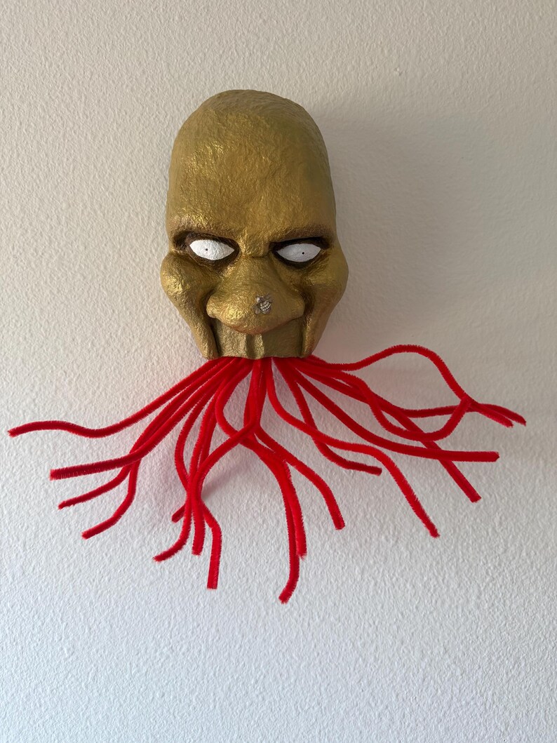 Puede incluir: Una m&aacute;scara dorada hecha a mano con ojos blancos y un dise&ntilde;o rojo en forma de tent&aacute;culos que se extiende desde la boca. La m&aacute;scara tiene una superficie texturizada y est&aacute; montada en una pared blanca, creando una exhibici&oacute;n art&iacute;stica &uacute;nica.