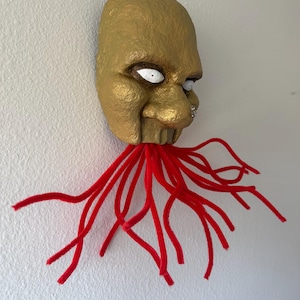 Puede incluir: Una m&aacute;scara dorada con ojos blancos y un piercing en la nariz, adornada con tent&aacute;culos de limpiapipas rojos. La m&aacute;scara tiene una superficie texturizada y est&aacute; montada en una pared blanca, creando una pieza decorativa &uacute;nica.