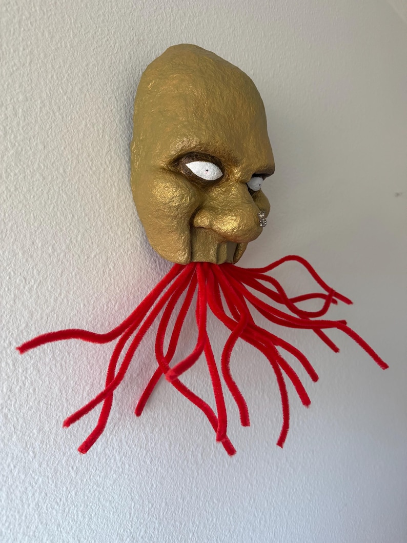 Puede incluir: Una m&aacute;scara dorada con ojos blancos y un piercing en la nariz, adornada con tent&aacute;culos de limpiapipas rojos. La m&aacute;scara tiene una superficie texturizada y est&aacute; montada en una pared blanca, creando una pieza decorativa &uacute;nica.
