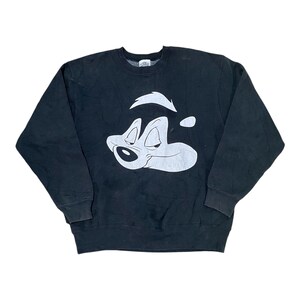 Puede incluir: Una sudadera negra de manga larga con cuello redondo que presenta un gran gráfico serigrafiado en blanco del personaje de dibujos animados Pepe Le Pew en la parte delantera. La prenda tiene cuello, puños y dobladillo acanalados.