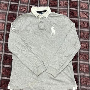 May include: A long-sleeve polo shirt in heather gray featuring a white collar and a white embroidered logo on the chest. The shirt has a buttoned placket, ribbed cuffs, and a small white graphic detail on the right sleeve near the shoulder area.