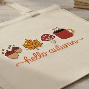 May include: Cream-colored tote bag with a fall-themed design. The design includes acorns, a maple leaf, a mushroom, and a steaming mug of coffee. The words "hello autumn" are written in cursive.