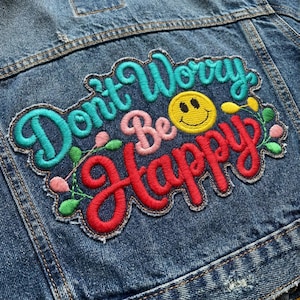 May include: Close-up of a denim jacket with an embroidered patch. The patch features the words "Don't Worry Be Happy" in teal, pink, and red, with a yellow smiley face and floral accents. The jacket is a medium wash blue.