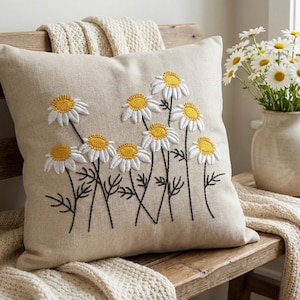 Puede incluir: Un cojín cuadrado beige con un diseño de margaritas bordadas. Las margaritas tienen pétalos blancos y centros amarillos, con tallos y hojas negras. El cojín está sobre un banco de madera con una manta color crema.
