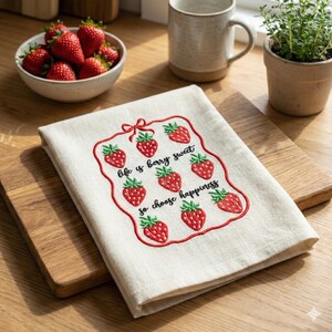 Puede incluir: Un paño de cocina color crema con un diseño de fresas bordadas. El diseño incluye fresas rojas y el texto "life is berry sweet so choose happiness" en escritura negra. El paño está doblado sobre una tabla de cortar de madera.