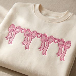 May include: Cream-colored sweatshirt with embroidered pink bows and hearts. The bows are arranged in a horizontal line across the chest. The sweatshirt has a crew neck and long sleeves.