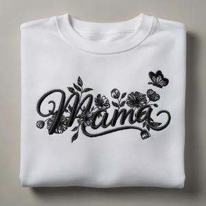 May include: White sweatshirt with the word "Mama" embroidered in black script, surrounded by floral and butterfly accents. The design is centered on the folded garment, creating a clean and stylish look.