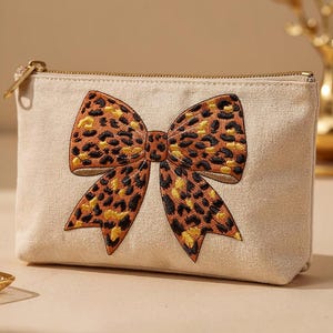 May include: A beige canvas cosmetic bag with a large embroidered bow. The bow features a leopard print pattern in shades of brown, black, and yellow. The bag has a gold zipper closure and is rectangular in shape.