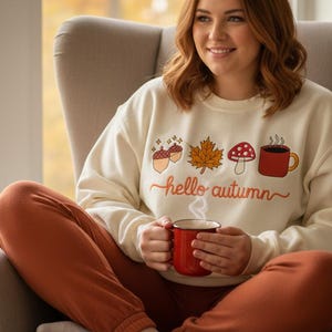 以下が含まれることがあります： 秋をテーマにしたグラフィックと「hello autumn」の文字が入ったクリーム色のスウェットシャツを着た人。赤いマグカップを持ち、灰色の肘掛け椅子に座っています。オレンジ色のパンツを履いています。