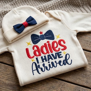 May include: Cream-colored baby outfit set with a matching beanie. The onesie features the embroidered text "Ladies I Have Arrived" in red and blue. The beanie has a navy blue bow tie with a red center.