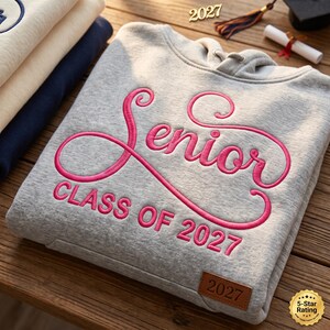 May include: A light gray hooded sweatshirt featuring the word Senior in large pink cursive embroidery, with the text CLASS OF 2027 underneath in pink block letters and a small brown rectangular leather patch embossed with 2027 at the bottom.