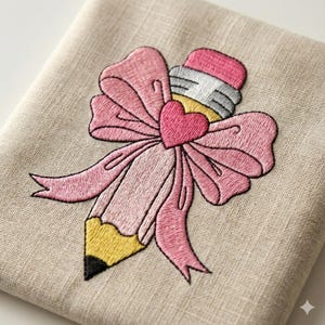 May include: Embroidered design of a pink pencil with a pink bow and a heart in the center. The pencil has a yellow body, a black tip, and a pink eraser. The design is on a beige linen fabric.