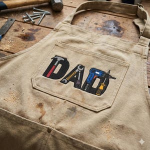 May include: Tan canvas apron with a pocket featuring the embroidered word "DAD" with tools incorporated into the letters. The tools include a hammer, wrench, drill, and screwdriver. The apron has a neck strap and waist ties.
