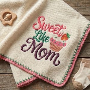Könnte beinhalten: Cremefarbene Babydecke mit der gestickten Aufschrift "Sweet Like Mom" und einem Cupcake mit Erdbeere. Die Decke hat einen rosa und grünen Rand. Ein hölzernes sternförmiges Rasselspielzeug und Babysocken sind ebenfalls zu sehen.