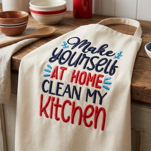 May include: Off-white kitchen apron with the embroidered text "Make yourself at home, clean my kitchen" in navy blue and red. The apron has a loop for hanging and is made of a natural-colored fabric. Kitchen and home decor.