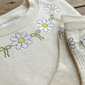 Op de afbeelding: Crèmekleurige sweatshirt- en sokkenset met geborduurd madeliefjeskettingontwerp. De madeliefjes hebben witte bloemblaadjes, gele centra en zijn verbonden door groene strikken. De sweatshirt heeft een ronde hals en de sokken zijn geribbeld.