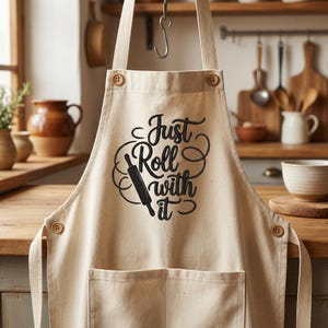 Könnte beinhalten: Beige Schürze mit dem Text "Just Roll with it" in schwarzer Schrift, mit einer Nudelholz-Grafik. Die Schürze hat eine Vordertasche und Holzknoten. Die Schürze hängt in einer Küchenumgebung.