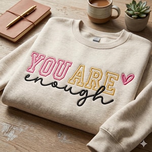 Może przedstawiać: Beżowa bluza z haftowanym napisem "YOU ARE enough" w kolorze różowym, żółtym i czarnym. Małe różowe serce znajduje się obok słowa "ARE". Bluza leży na drewnianej powierzchni z notatnikiem, długopisem, filiżanką kawy i rośliną.