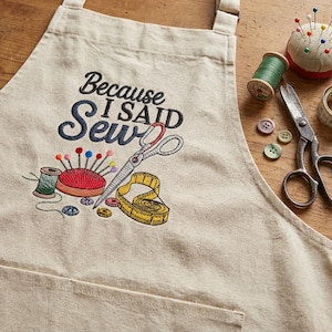 A beige apron with a humorous embroidered phrase "Because I Said Sew" is displayed on a wooden surface, accompanied by sewing tools such as scissors and pins.