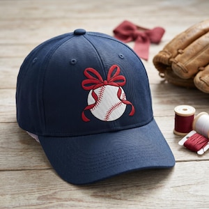 Op de afbeelding: Marineblauwe baseballpet met een geborduurd honkbalontwerp. De honkbal is wit met rode stiksels en een rode strik. De pet ligt op een houten oppervlak.