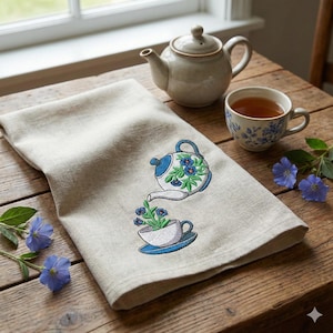 May include: A beige linen tea towel featuring an embroidered teapot and teacup design. The teapot is pouring blue flowers into the teacup. A ceramic teapot and teacup with tea are on a wooden table, with blue flowers.
